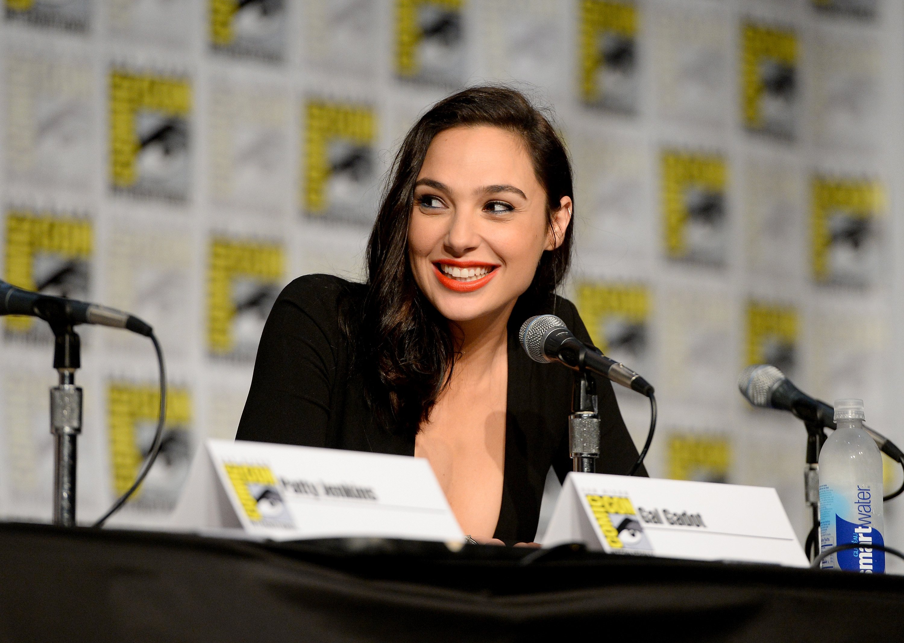 Gal Gadot at San Diego Comic Con, smiling and speaking into a microphone while sitting at a panel discussion. Gal Gadot at San Diego Comic Con, smiling and speaking into a microphone while sitting at a panel discussion.