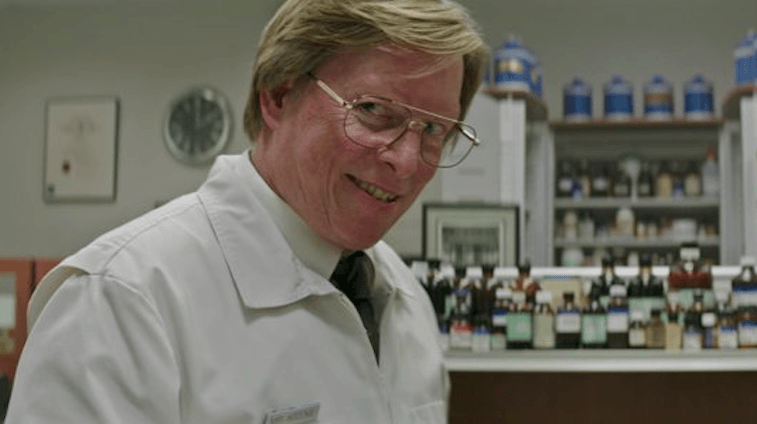 A pharmacist wears glasses and stands in front of a shelf filled with medicine A pharmacist wears glasses and stands in front of a shelf filled with medicine