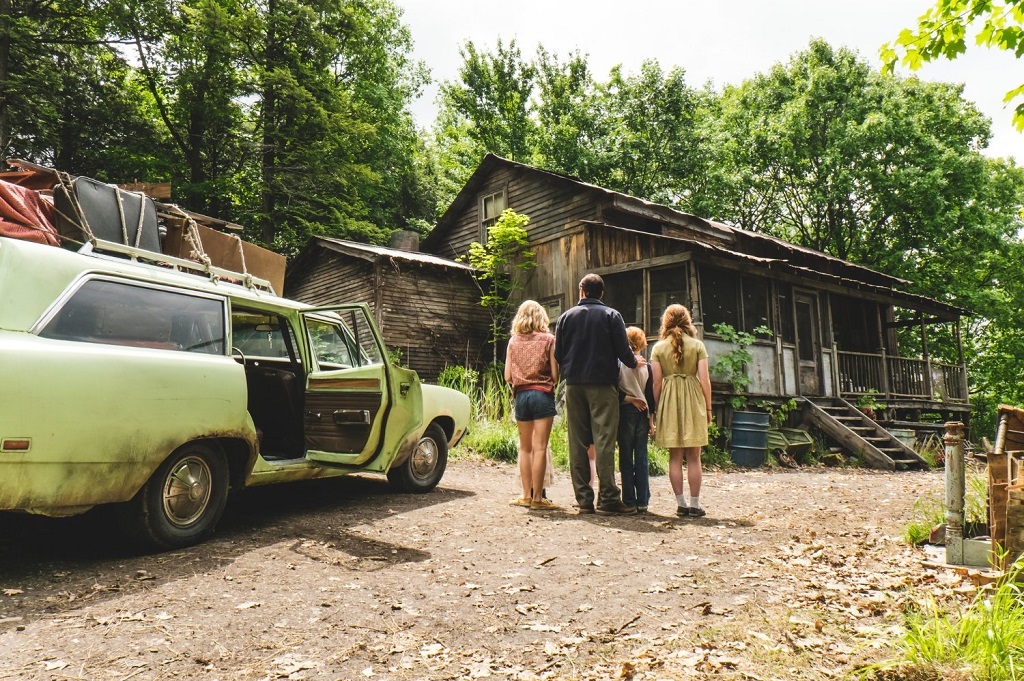 A family stands next to their green car in the woods looking at a rundown cabin