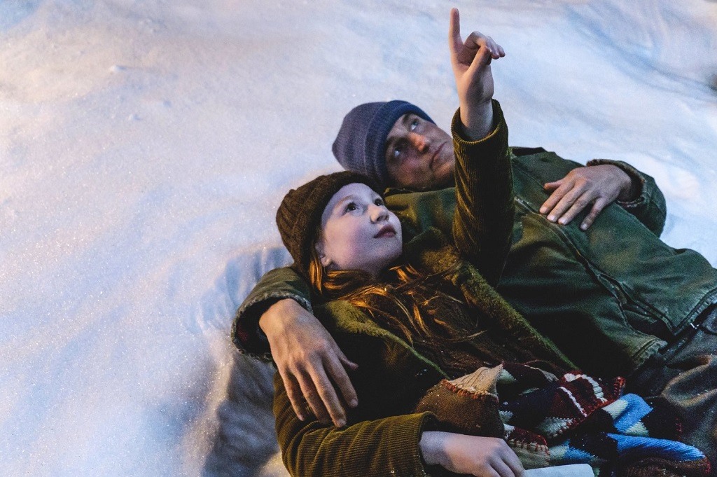 A child and her father lay in the snow as she points up at the sky