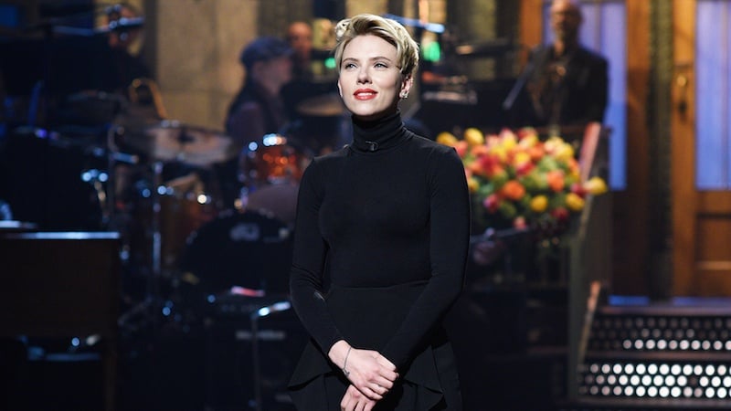 Scarlett Johansson stands on the sNL stage in a black turtleneck with her hands crossed in front of her