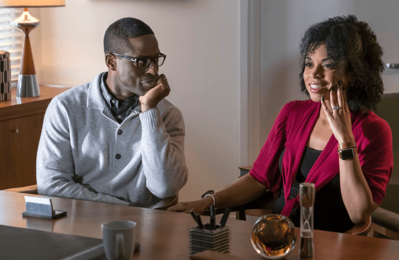 Randall stares at Beth as she smiles and looks ahead across a desk. Randall stares at Beth as she smiles and looks ahead across a desk.