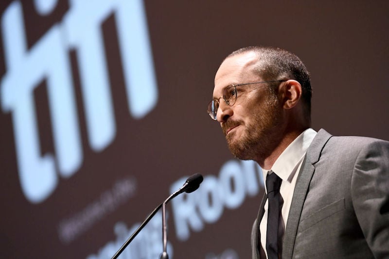 Darren Aronofsky speaks front on a microphone while wearing a gray suit.