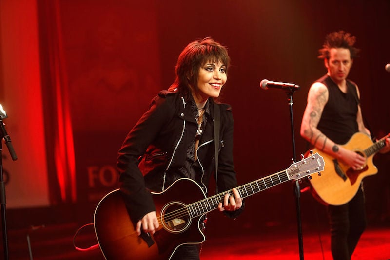 Joan Jett performing on stage with a guitar in front of a microphone.