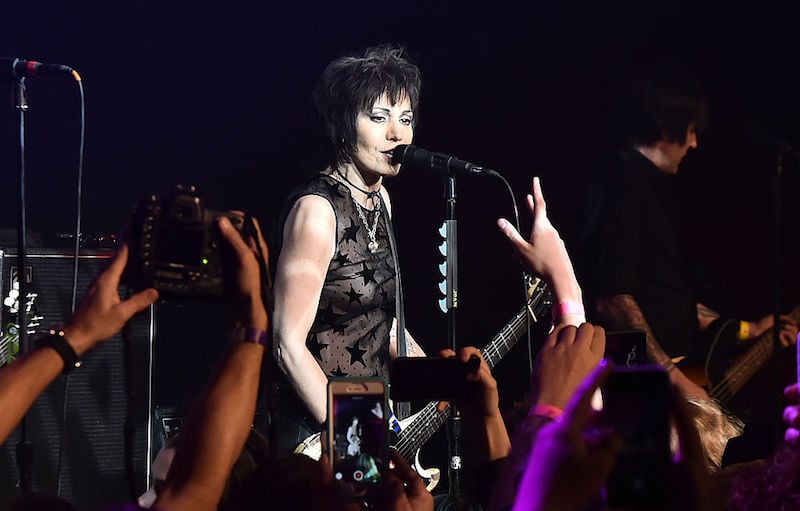 Joan Jett singing into a microphone as fans in the audience take photos.
