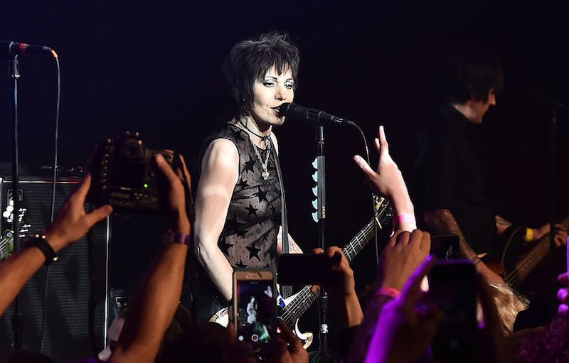 Joan Jett singing into a microphone as fans in the audience take photos. Joan Jett singing into a microphone as fans in the audience take photos.