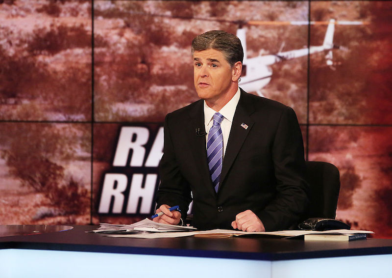 Sean Hannity sitting behind a news desk while holding a pen and looking straight ahead. 
