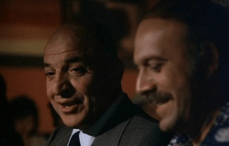 Telly Savalas turning to the side and talking to another person. 