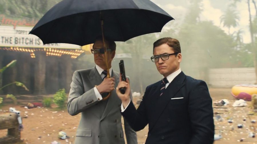 Colin Firth and Taron Egerton stand next to each other while holding a gun and umbrella