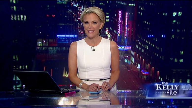 Megyn Kelly sitting at a news desk with a phone and laptop in front of her. 
