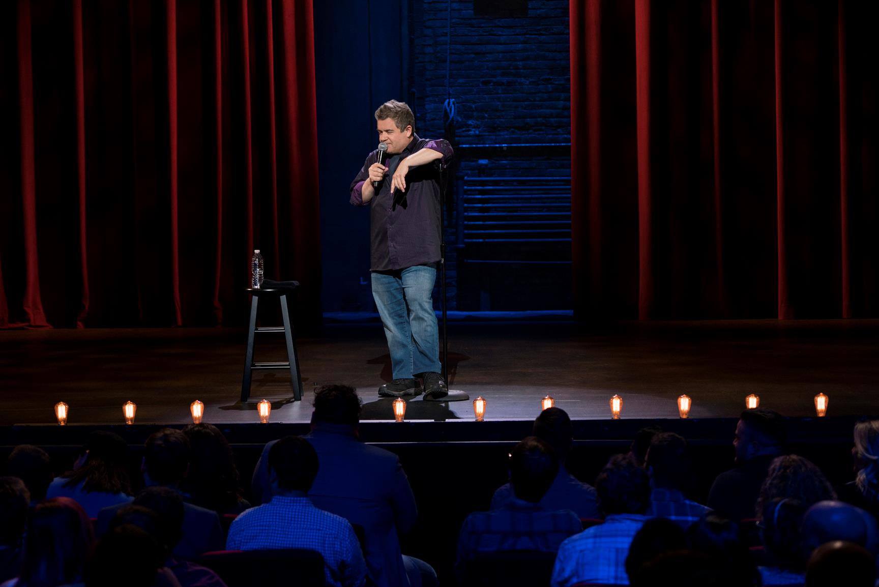 Patton Oswalt stands on stage holding a microphone