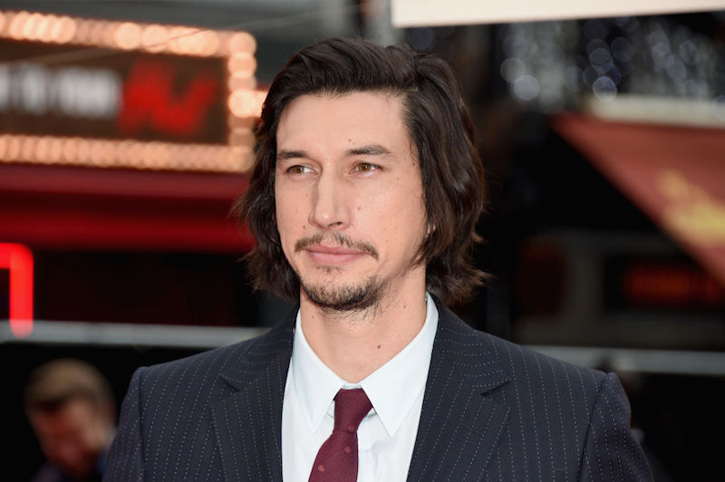 Adam Driver posing for photos on a red carpet in a dark suit and red tie. 