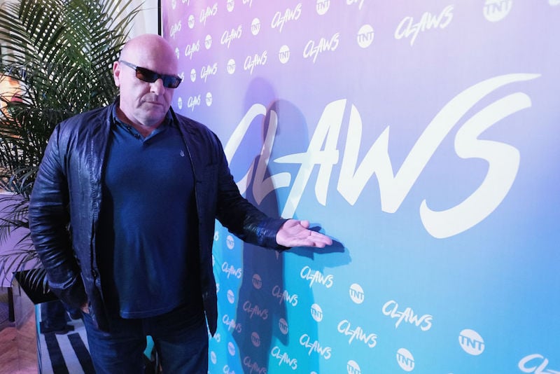 Dean Norris pointing at a sign at a red carpet event while wearing shades and a leather jacket.