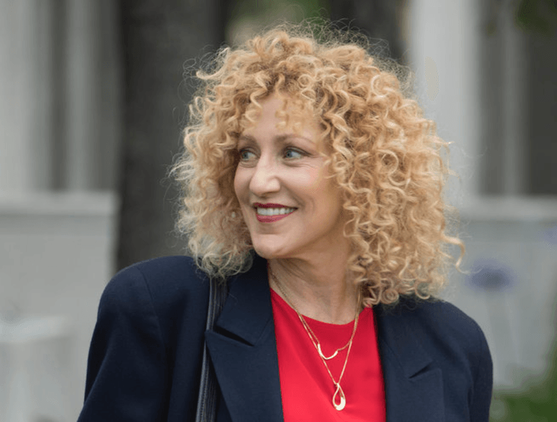 Edie Falco smiling and looking backwards while wearing a red shirt and black blazer. Edie Falco smiling and looking backwards while wearing a red shirt and black blazer.
