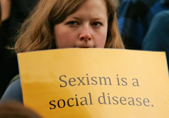 a woman holds a yellow sign reading sexism is a social disease