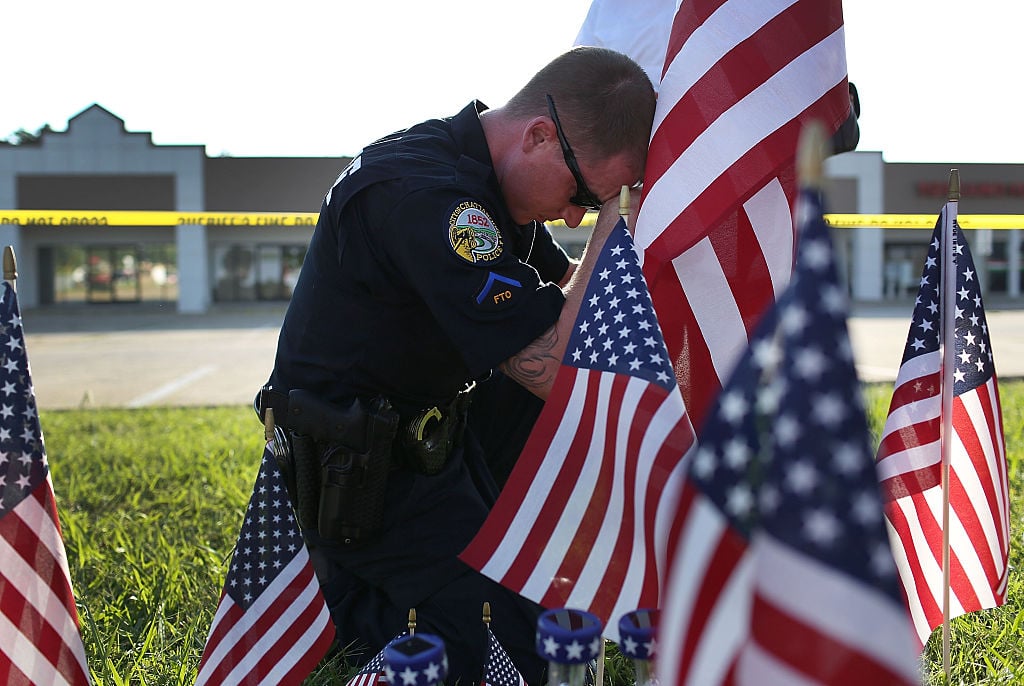 An officer on his knees places a flagamong many flags at the site of a memorial
