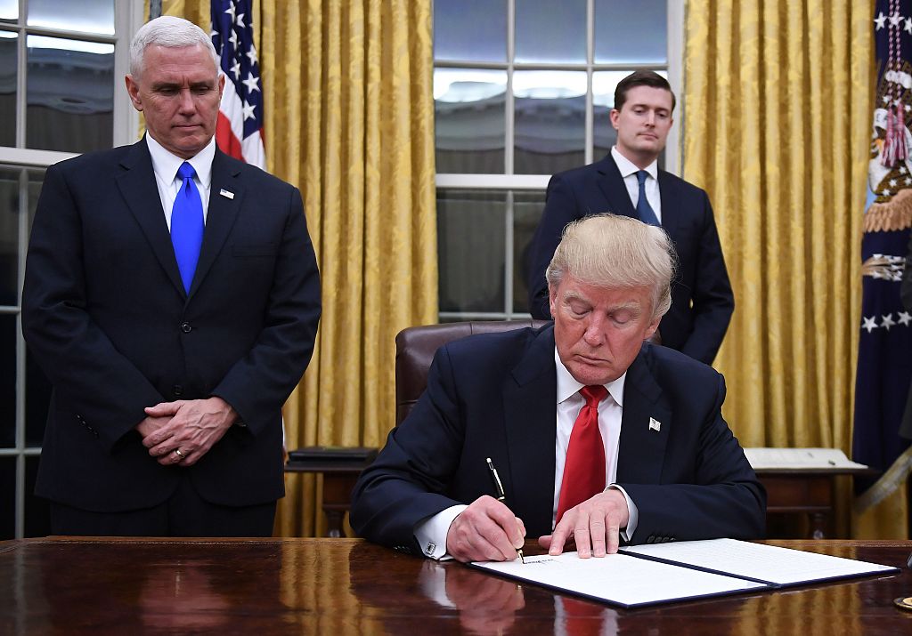 Donald Trump wearing a red tie and a dark suit signs a paper sitting at his desk