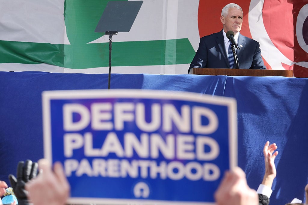 Mike Pence speaking with a blue and white defund planned parenthood sign in the foreground Mike Pence speaking with a blue and white defund planned parenthood sign in the foreground