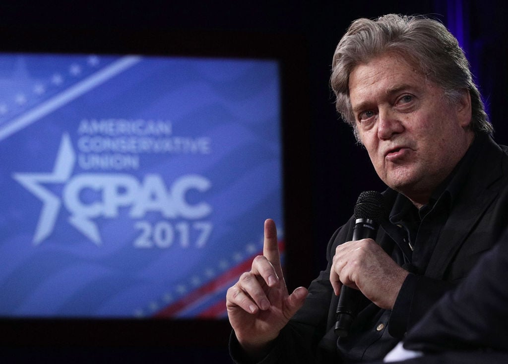 Steve Bannon on the right, looking left, in front of a blue and white poster Steve Bannon on the right, looking left, in front of a blue and white poster