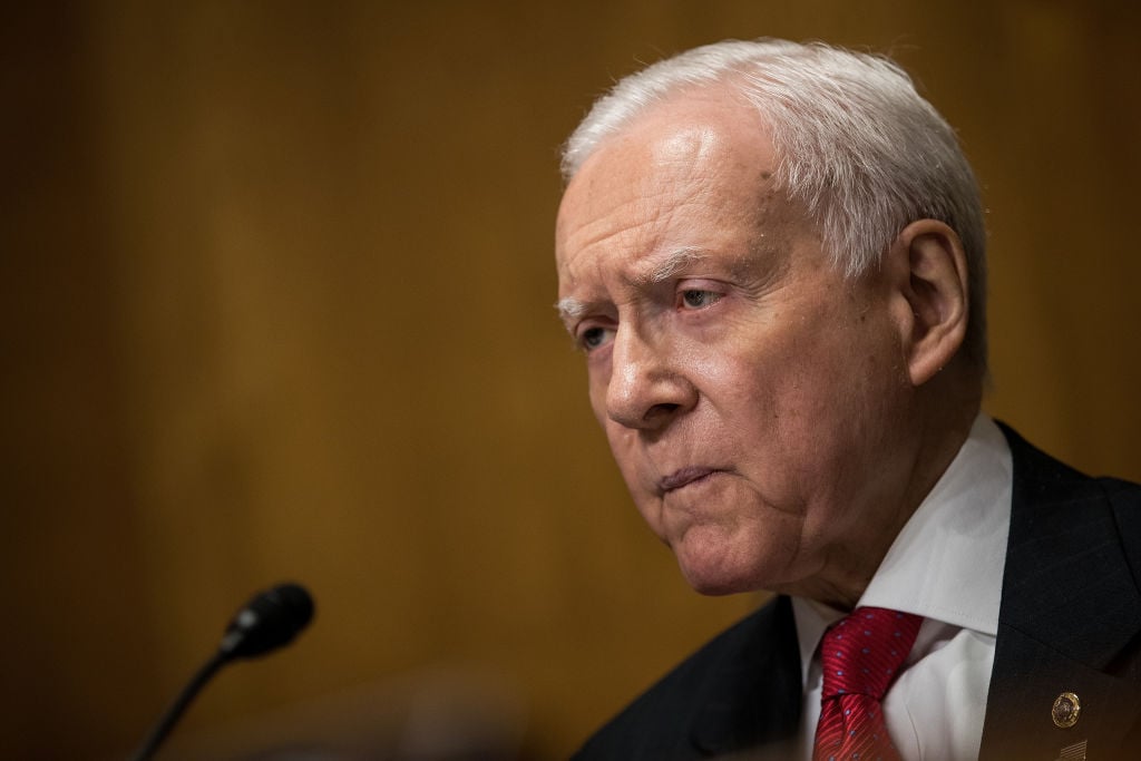Orrin Hatch, facing left with a microphone against a wood background Orrin Hatch, facing left with a microphone against a wood background