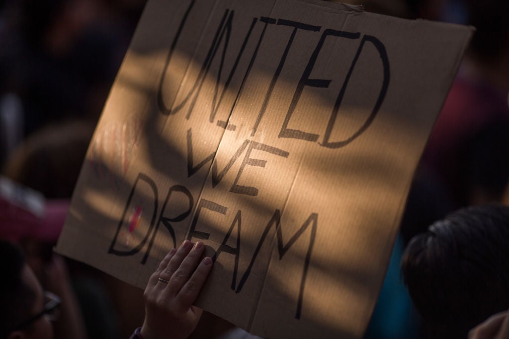 a hand holding up a cardboard sign with United We Dream on it in black sharpie a hand holding up a cardboard sign with United We Dream on it in black sharpie