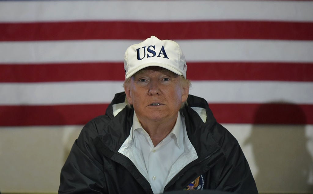 donald trump wearing a USA hat and a windbreaker against an american flag