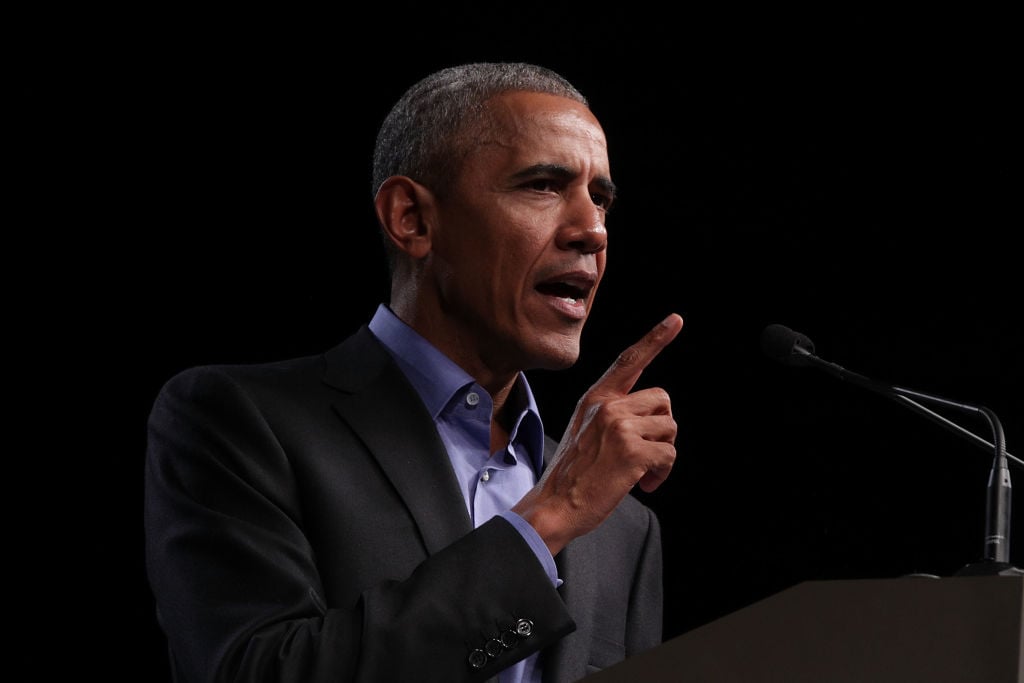 Barack Obama speaking in a dark suit into a microphone.