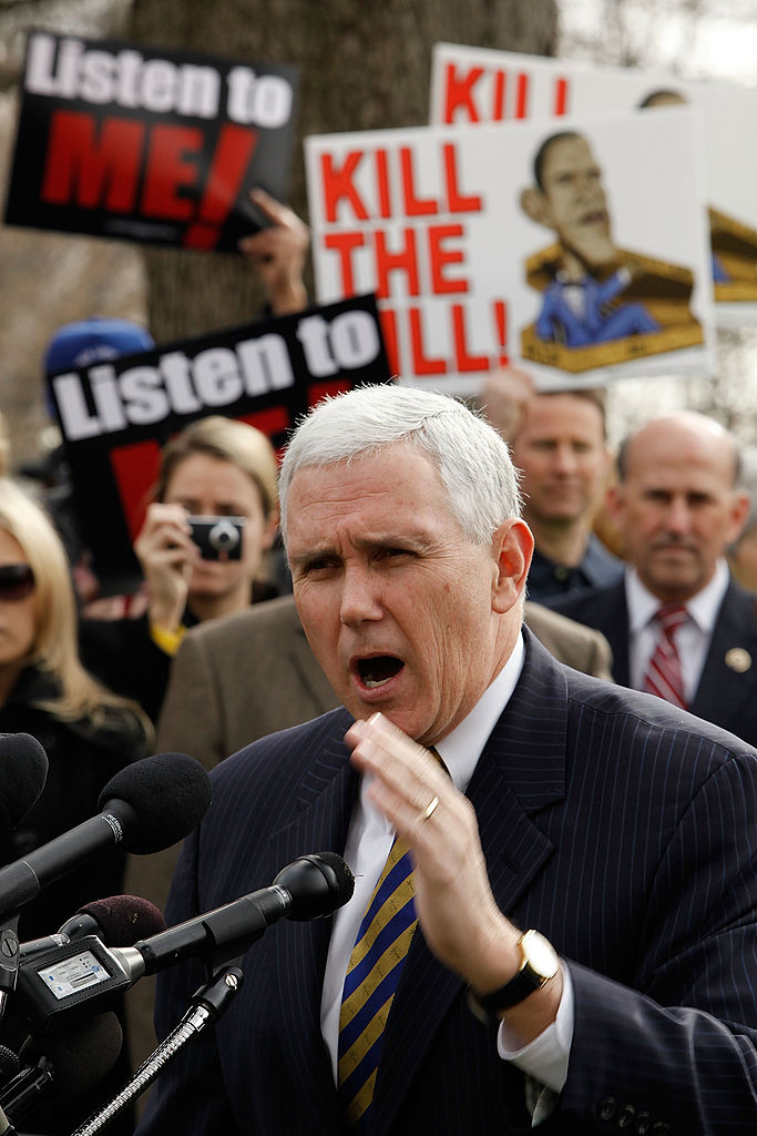 Mike Pence in a dark suit speaking against Tea party protest signs
