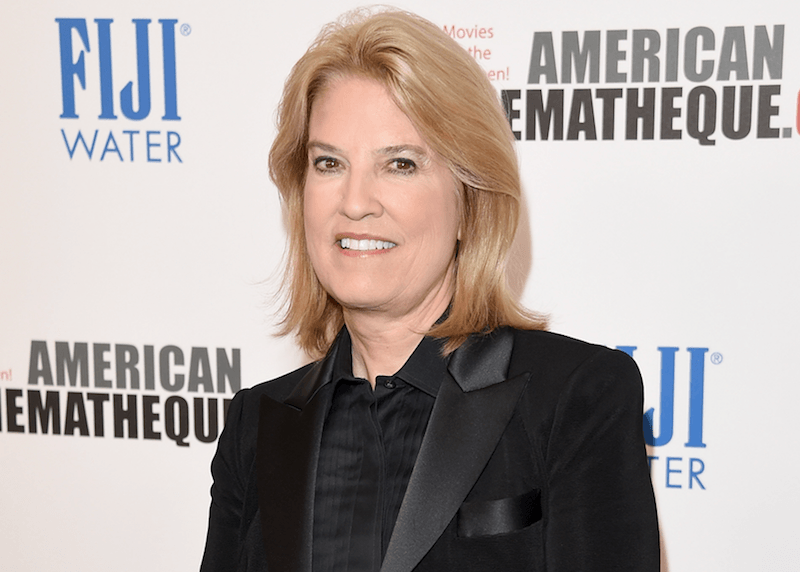 Greta Van Susteren standing in a black tuxedo on a red carpet.
