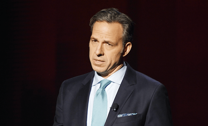 Jake Tapper stands in a dark suit and light blue tie behind a dark curtain. Jake Tapper stands in a dark suit and light blue tie behind a dark curtain.