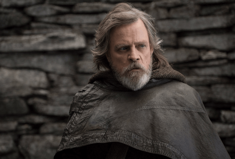 Luke Skywalker stands in a brown cloth in front of a brick wall as he looks towards the side. 