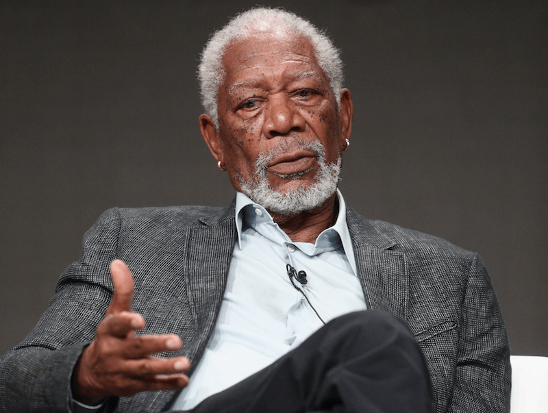 Morgan Freeman sitting and speaking while on a stage. Morgan Freeman sitting and speaking while on a stage.