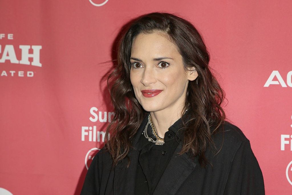 Actress Winona Ryder smiling in front of a pink backdrop at a red carpet event.