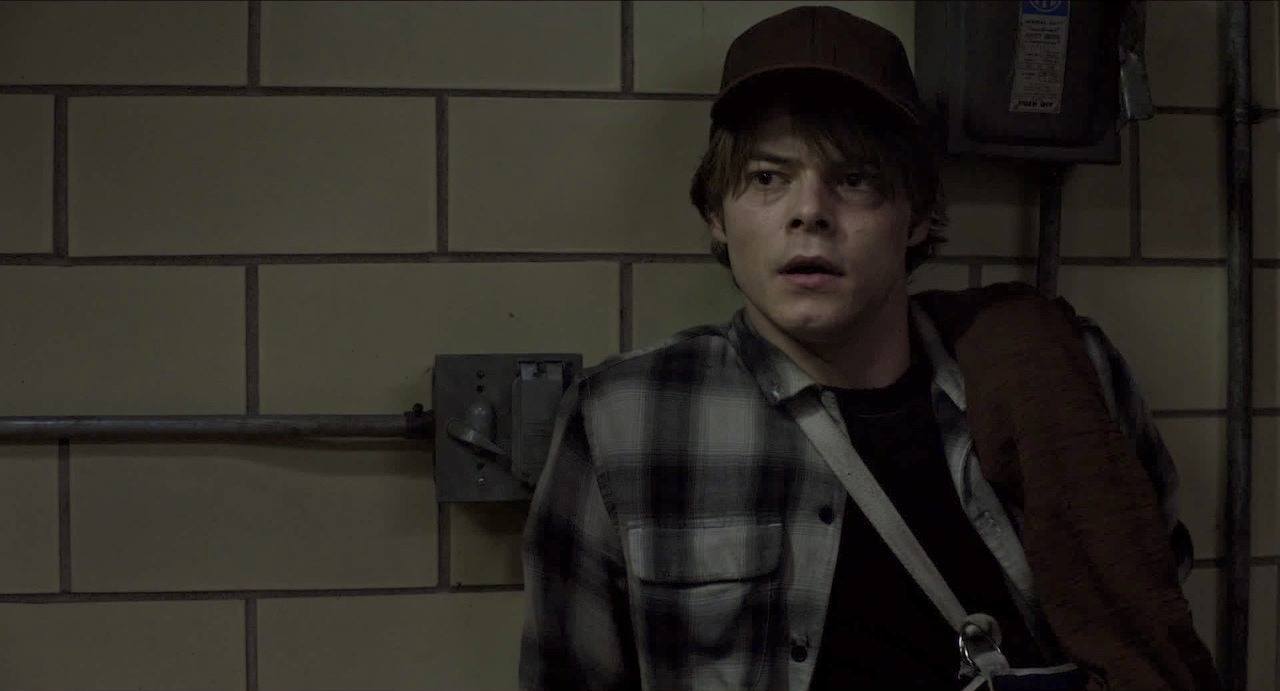 Charlie Heaton's Cannonball leans against a tile wall while wearing a flannel shirt 