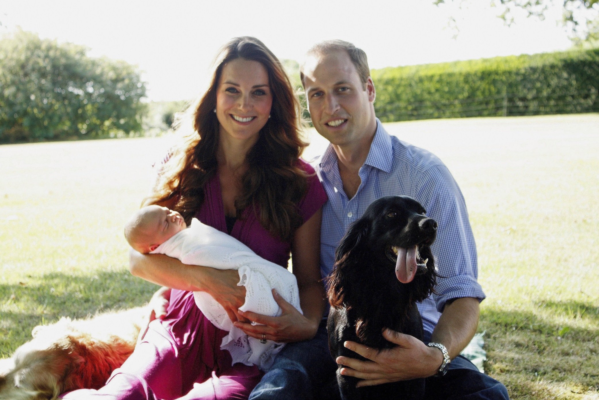 The royal family sits outside in a garden with their baby and dog. 