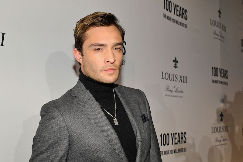 Ed Westwick posing on a red carpet in a black turtle neck sweater and gray blazer. 