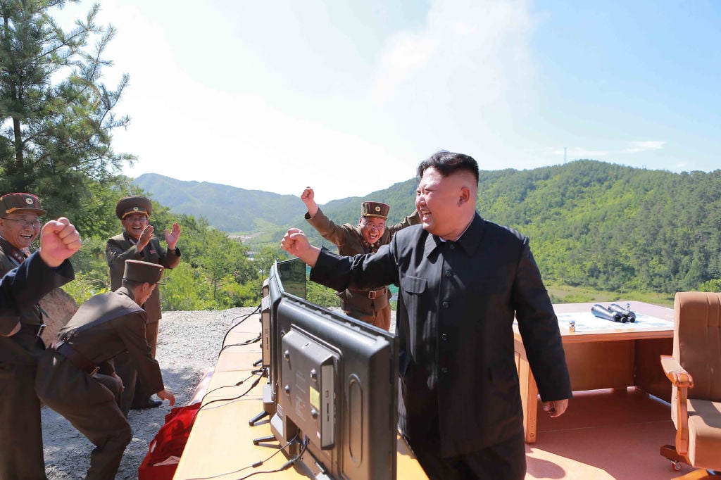 Kim Jong Un stands in celebration with a bunch of his generals in front of monitors on top of a mountain on a sunny day.