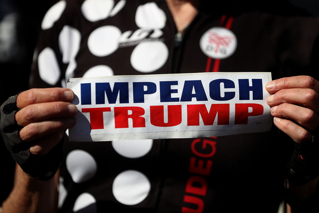 Normal sized hands hold a bumper sticker that reads "Impeach Trump" in blue and red lettering
