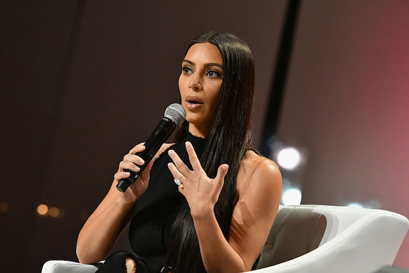 Kim Kardashian speaks into a microphone while sitting on a white chair on stage.