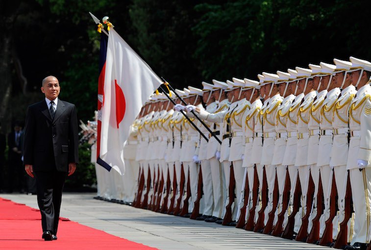 King Norodom Sihamoni of Cambodia walks on a red carpet alongside military men.