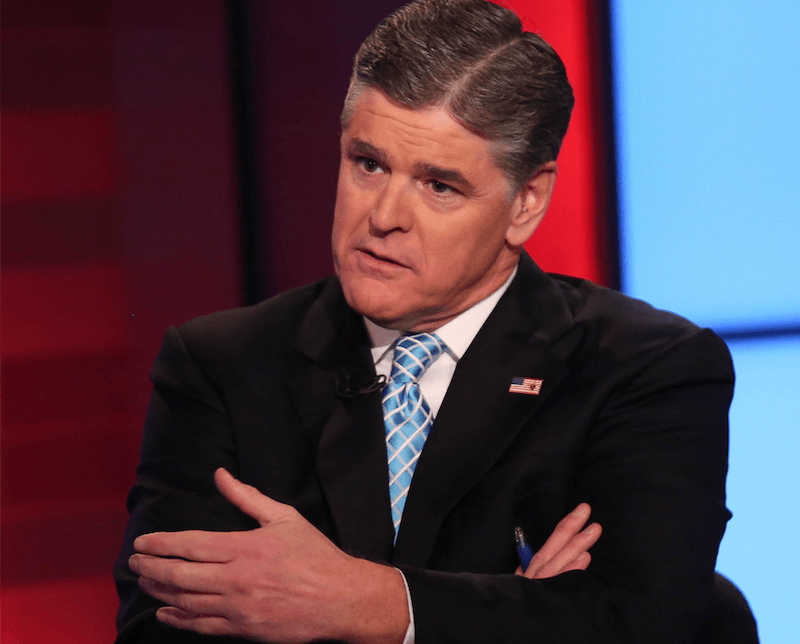 Sean Hannity sits while crossing his arms at his desk. 