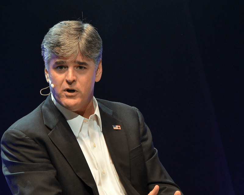 Sean Hannity speaks while wearing a gray suit and an earpiece. 