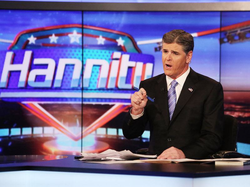 Sean Hannity holding a pen while sitting behind a news desk. 
