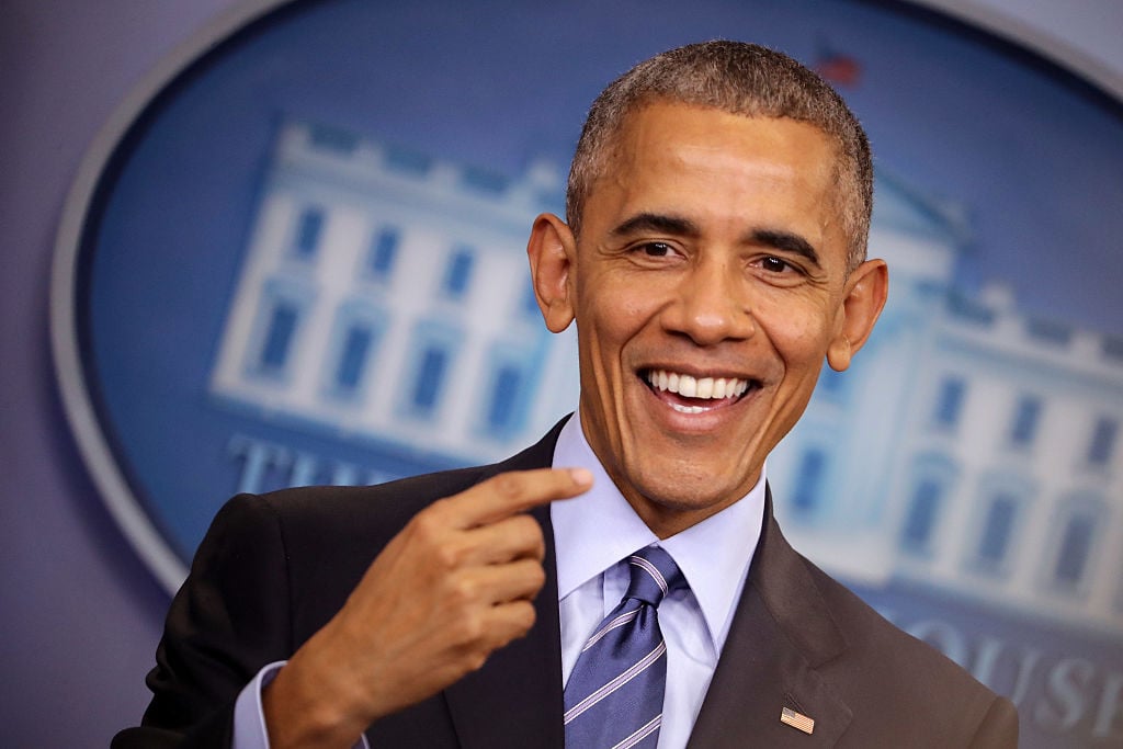 Barack Obama pointing at his own face in a dark suit and blue striped tie