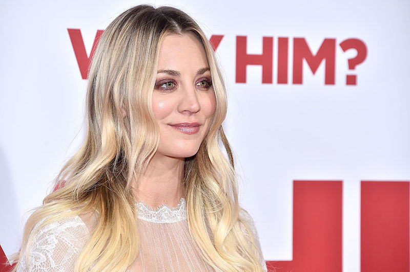 Kaley Cuoco smiles and poses on a red carpet wearing a white lace dress.