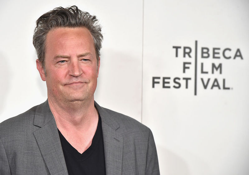 Matthew Perry posing in a grey suit and black shirt. 