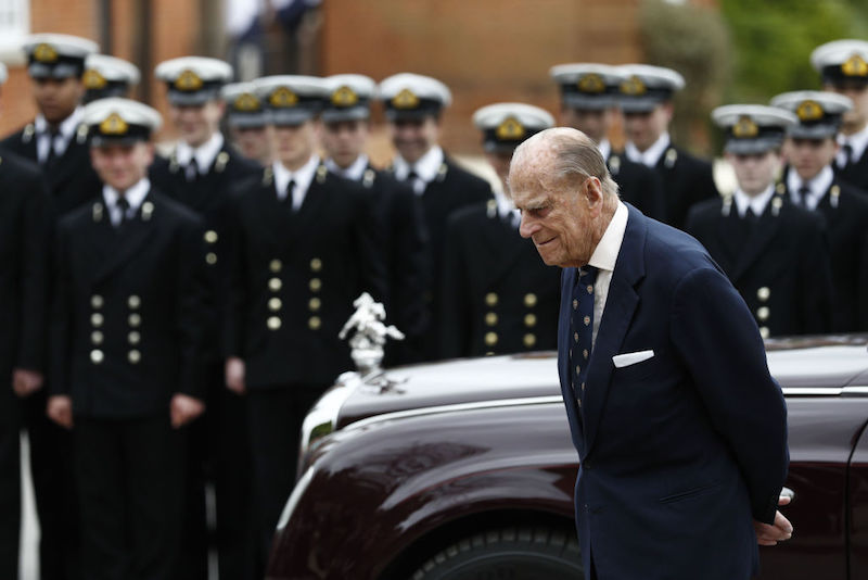 Prince Phillip stands in front of a naval army and shiny car. 