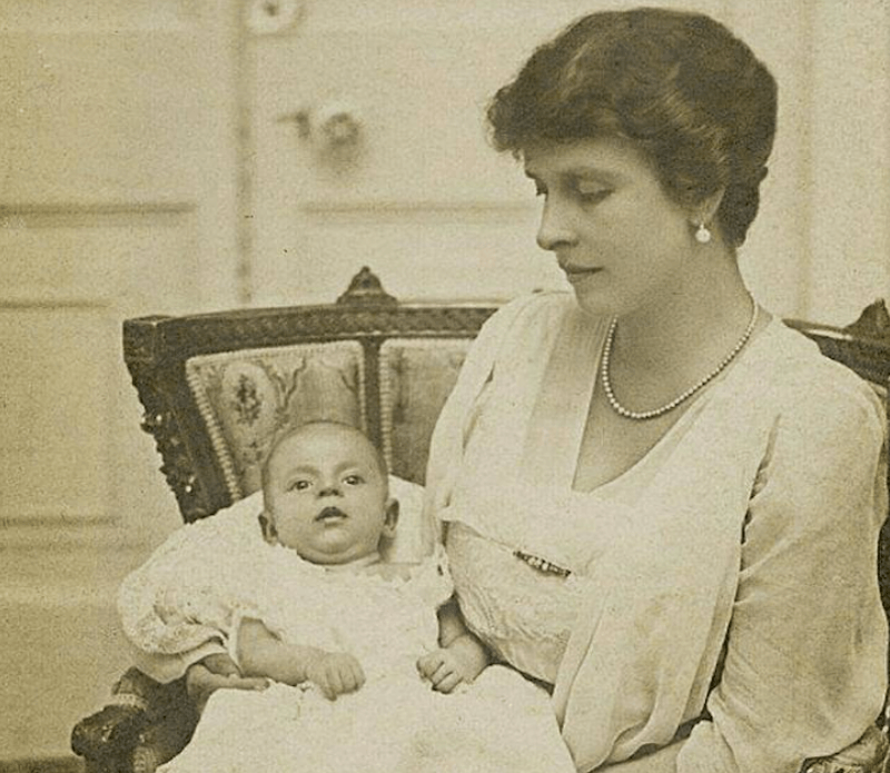 Princess Alice holding her baby while wearing a suit and jewelry. 