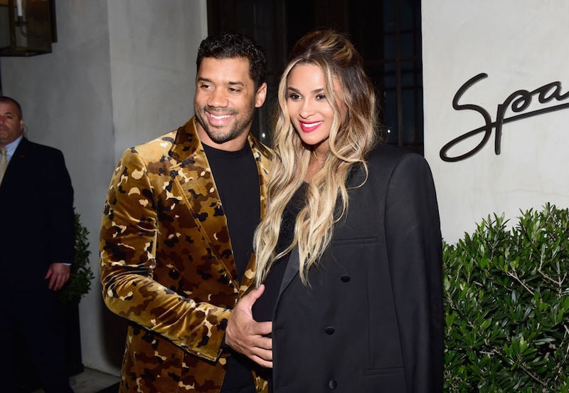 Russell Wilson places his hand on Ciara's baby bump as they smile and pose on a red carpet.