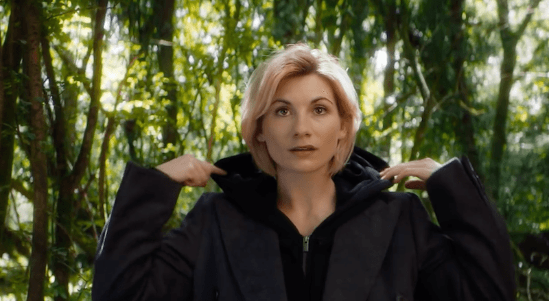 The Doctor stands in a forest wearing black. 